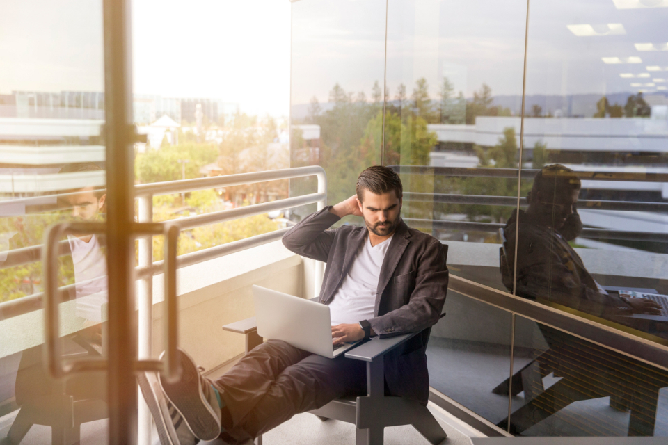 A man at a desk in an office, looking confused while working at a laptop.